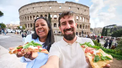Un bărbat în tricou polo alb și o femeie într-o cămașă albastră zâmbesc la cameră ținând câte o felie de pizza în mână și au pe fundal Colosseumul din Roma