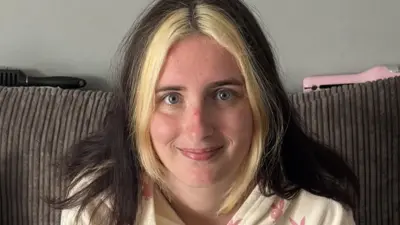A close-up photo of Kailee who looks into the camera with a smile, she is sat on her sofa at home. She has dark hair down over her shoulders, apart from the front which is dyed blonde.