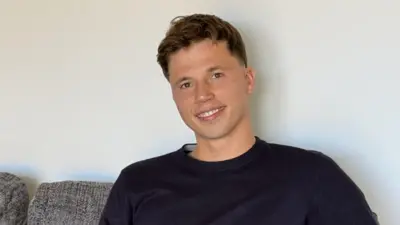 Freddie Woodward: un joven de pelo corto y oscuro está sentado en un sofá gris, sonriendo a la cámara. Tiene el pelo corto castaño y lleva un jersey azul marino.