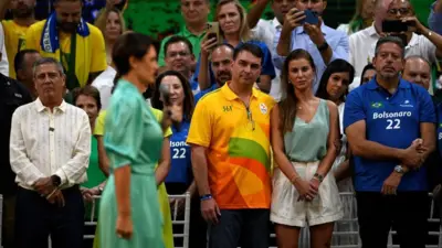 Michelle aparece em primeiro plano, desfocada, em vestido verde e falando ao microfone em um comício eleitoral de 2022. Ao fundo, Flávio e sua esposa observam Michelle ao lado de outras pessoas
