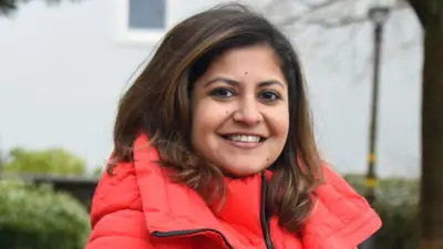 A woman with shoulder-length brown hair smiles. She is wearing a large salmon-coloured puffer jacket.