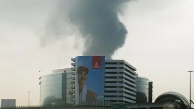 Smoke rising from an area near Dubai International Airport is seen through a vehicle’s windshield after a drone attack hit a fuel tank. In the foreground, an Emirates poster portraying a female flight attendant covers almost the entire façade of a building