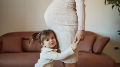 Küçük bir kız çocuğu sadece karnı görünen hamile annesinin ellerinden tutarak ona sarılmış halde ayakta durmuş kameraya bakıyor