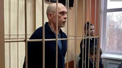 Andrey Perlov standing behind bars in court, wearing a dark blue top, there are two guards nearby