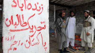 سه مرد با لباس محلی پاکستانی ایستاده‌اند که یکی از آنها سلاح ای‌کی روی دوشش دارد و یکی دیگر جلیقه نظامی پوشیده است