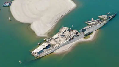 Destroços de um navio encalhado em um banco de areia, com pessoas observando