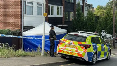 A police officer guarding the cordon at the McCullough property in Pump Hill. They have a police car in front of them and to their side is a blue sided police tent with a white top. 