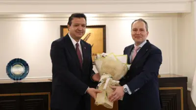 İki takım elbiseli erkek, bir demek çiçeği tutuyorlar. Arkada Yeniden Refah Partisi logosu var