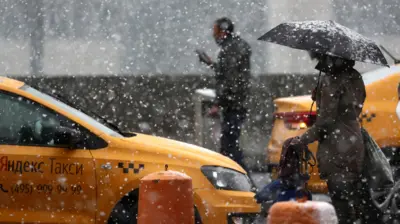 Падает снег. На переднем плате желтая машина "Яндекс Такси". Справа от нее человек под зонтиком.