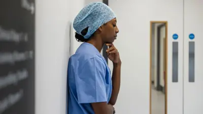 An NHS hospital worker wearing scrubs and a hair net stands against a wall inside a hospital in Newcastle while looking pensive. Stock photo illustration.