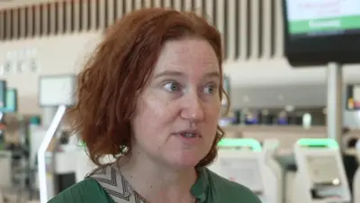 Kate White, que tem cabelo ruivo curto, fala em uma entrevista na TV em frente aos balcões de check-in no saguão de embarque do Aeroporto de Manchester.