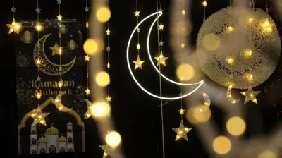 Golden lights and decorations for Ramadan in bazaar in Sana'a, Yemen. There is a cescent Moon in the background above an image of a mosque with the words Ramadan Mubarak written above it. The outline of a white crescent Moon is in the foreground. Some golden stars are hanging around the area.