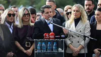 Özer resmi giysili ve bir kürsüde mikrofonlara konuşşuyor sağında ve solunda ikişer kadın arkasında da bir grup insan var. 