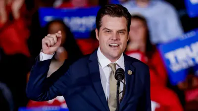 Matt Gaetz stands with one arm raised in the air, speaking before a crowd of people holding red and blue signs. He has on a blue suit and gold tie