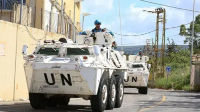 2台の大型の白い戦車が曲がりくねった道を走行している。戦士の正面には「UN
（国連）」と大きく書かれている。明るい青色のヘルメットをかぶった国連要員が各車両の上に立っている