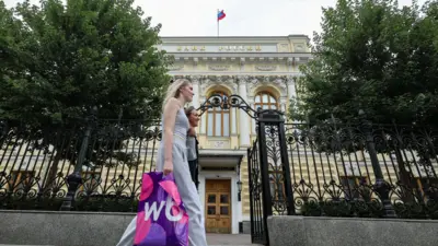 Dwie kobiety przechodzą obok bladożółtego budynku z rosyjskim napisem „Bank Rosji” i rosyjską flagą w kolorach białym, niebieskim i czerwonym.
