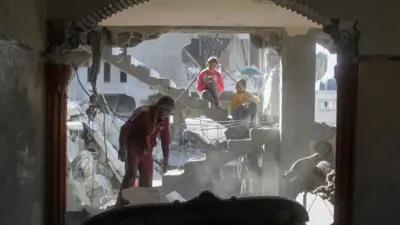 A Palestinian man, wearing a red tracksuit, inspects rubble left from a house hit by a strike, as two young girls sit on a staircase