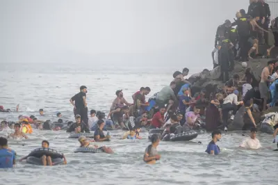 Dezenas de imigrantes de boia ou a nado chegam pelo mar e escalam rochas à beira de um cercado