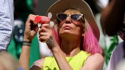 A person raises a fan to their face while wearing a sunhat on a hot, sunny day at Wimbledon on 10 July