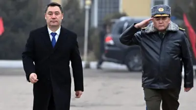 Қирғизистон президенти Садир Жапаров ва Миллий хавфсизлик давлат қўмитасининг собиқ раиси Қамчибек Ташиев бирга кетмоқда. Жапаров қора палто, оқ кўйлак ва кўк галстукда одимламоқда. Унинг ёнида Ташиев ҳарбий қишки формада, ҳарбийларча қўлини чеккасига қўйиб бормоқда.