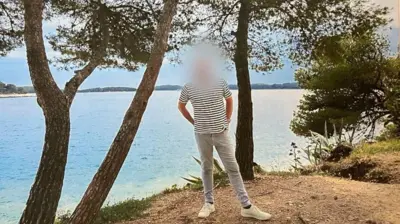 Image shows a man in pale jeans and a striped t shirt. His face has been blurred out. He is stood in front of a lake, surrounded by trees. The sky is blue and he has his hands in his back pockets, looking relaxed.