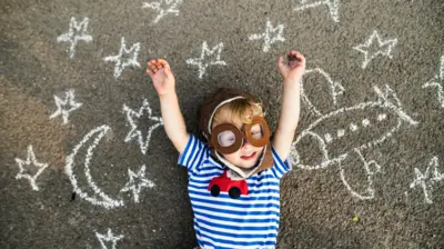 Menino sorridente usando chapéu de piloto e óculos de proteção, deitada no asfalto pintado com desenhos de avião, lua e estrelas.