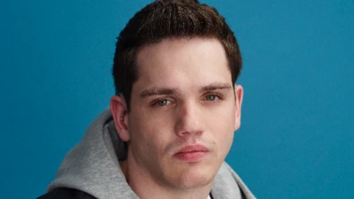 Elliott Caswell looks directly at the camera, wearing a grey hooded top and a black bodywarmer and white t-shirt. He has a serious expression and is pouting slightly. 