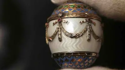 A Christie&#x27;s employee holds up a Fabergé enamel and two-colour gold egg bonbonniere in front of a portrait of King George I of the Hellenes. The egg has gold detailing on it, with a white centre, and purple and green detailing at the top and bottom.