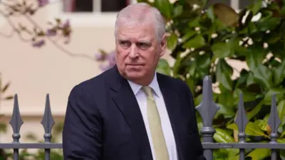 黒いブレザーに白いシャツ、金色のネクタイを着けたアンドリュー元王子が、グレーの金属製フェンスの前に立ち、厳しい表情で右の方を見ている。フェンスの奥に立つ木には、緑の葉が生い茂っている