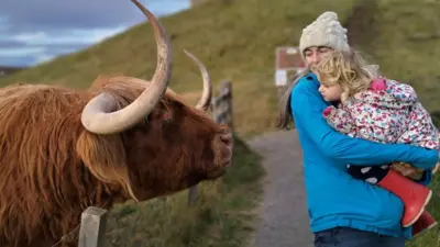 A large cow with horns leans over a fence, looking at a woman and a young girl