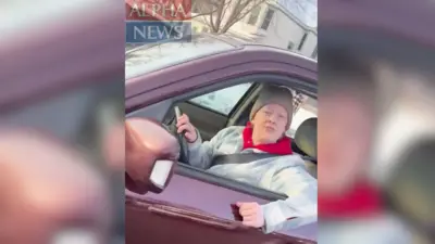 Image shows a woman wearing a blue and white jacket, a red hooded jumper and a beige hat. She is sitting in a red vehicle with the window open and her arm resting on the window frame