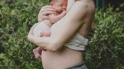 Kobieta tuli do piersi noworodka. Na zdjęciu widoczny jest jedynie jej tułów i ramiona. W tle zielony krzew lub żywopłot. 
