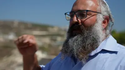 Yehuda Shimon, a large, bearded man wearing glasses, gestures with his hand