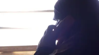 مرجان که حجاب و ماسک صورت سیاه دارد، در مقابل کلکین ایستاده و تلفون سرخ رنگ در دست دارد و در حال صحبت است.