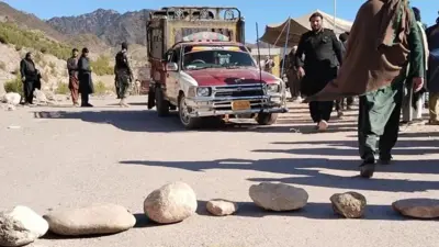 مسیری مسدود شده در تیراه سنگ های در وسط جاده گذاشته شده و یک موتر باربری متوقف شده است. چند نفر دورو بر آن هستند. در پس‌زمینه کوه‌های به چشم می‌خورند.