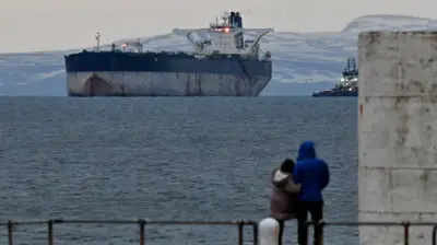 Пара в теплых куртках рассматривает с берега танкер Marinera в холодном шотландском заливе на фоне заснеженных холмов