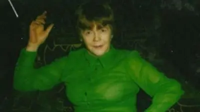 A young Mary McLaughlin poses while she dances, one hand on her hip, one in the air. She has short brown hair and is wearing a bright green chiffon shirt. She looks to be in a bar, with a dark background.