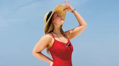 Hannah Howie, who has long, blonde hair and is wearing a red dress with white spots, strikes  a pose against a clear blue sky. Her right hand is on her hip and he left hand is tipping the top of her straw hat.