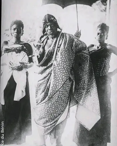 Photo archives du roi Behanzin portant un grand pagne autour de la taille avec une longue pipe entre les lèvres. Derrière lui se trouvent deux femmes dont l'une tient une parapluie pour protéger le roi.