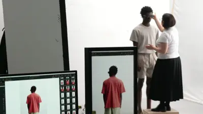 Male model standing wearing a light-coloured T-shirt and shorts with a woman standing on a block in front of him adjusting his sunglasses. Computer screens in the foreground show pictures of the same model taken from behind with him wearing a red T-shirt.