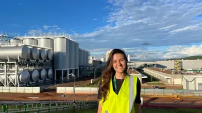 Letícia Lemos Martins veste um colete amarelo refletivo e sorri, com a fábrica da Heineken em Passos, Minas Gerais, ao fundo. Ela tem cabelos ondulados compridos e veste uma camiseta preta por debaixo do colete