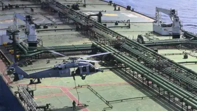 Sebuah helikopter militer berwarna abu-abu terbang di atas sebuah kapal tanker minyak, seorang prajurit meluncur turun dengan tali menuju dek atas sementara prajurit berseragam lainnya menunggu. Dek dipenuhi peralatan pengangkutan minyak, termasuk pipa-pipa hijau.