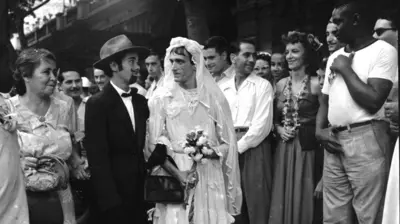 Fotografia em preto e branco tirada no carnaval de rua do Rio de 1954 mostra ao centro um casal de noivos, com o homem fantansiado de noiva e a mulher, de noivo, e várias pessoas sorridentes em volta