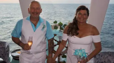A couple on a cruise ship smiling. The man (left) wears a blue shirt and white waistcoat. The woman (right) wears a white dress and carries blue flowers. She has curly brown hair. The sea is visible behind them.