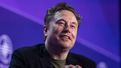 Elon Musk patrzy z uśmiechem w stronę kamery, siedząc na scenie na fioletowym tle