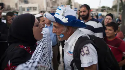 Uma mulher palestina usando um lenço na cabeça e um homem israelense usando um chapéu azul e branco com a bandeira israelense gritam um com o outrowelcome fortune slotJerusalém