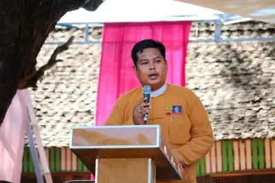 တိုင်းလွှတ်တော်အမတ်အဖြစ်မြင်ရတဲ့ ကိုဝဏ္ဏအောင် (ပုံဟောင်း)ကို ပင်နီတိုက်ပုံဝတ်ပြီး စင်မြင့်ထက် စကားပြောနေပုံမြင်တွေ့ရ၊ အနောက်မှာ သက်ကယ်မိုး အဆောက်အဦ အမိုးကို မြင်တွေ့ရ 