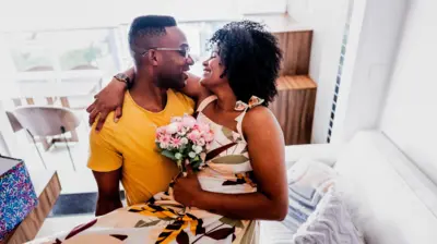 Casal sorridente abraçado ao lado de um sofá, olhando um para o outro; ela segura um pequeno buquê de flores em uma sala iluminada.