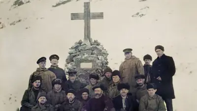 A group of 16 men from the crew of the Quest exploration ship, standing and sitting in front of a memorial cross to Sir Ernest Shackleton