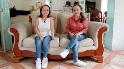 Maria Luisa, kız kardeşi María del Cisne ile krem rengi bir kanepede oturuyor. Maria Luisa'nın omuz hizasında kahverengi saçları var ve beyaz bir atlet, kot pantolon ve spor ayakkabı giymiş, gülümsüyor. Maria del Cisne'nin daha uzun koyu saçları var, ön kısmı arkaya doğru toplanmış, beyaz puantiyeli kırmızı bir tişört giymiş ve o da kot pantolon ve beyaz spor ayakkabı giyiyor.
